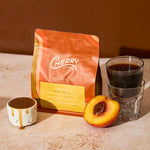 Cherry coffee cold brew retail bag with a small cup of caramel, cold brew, and half of a peach.