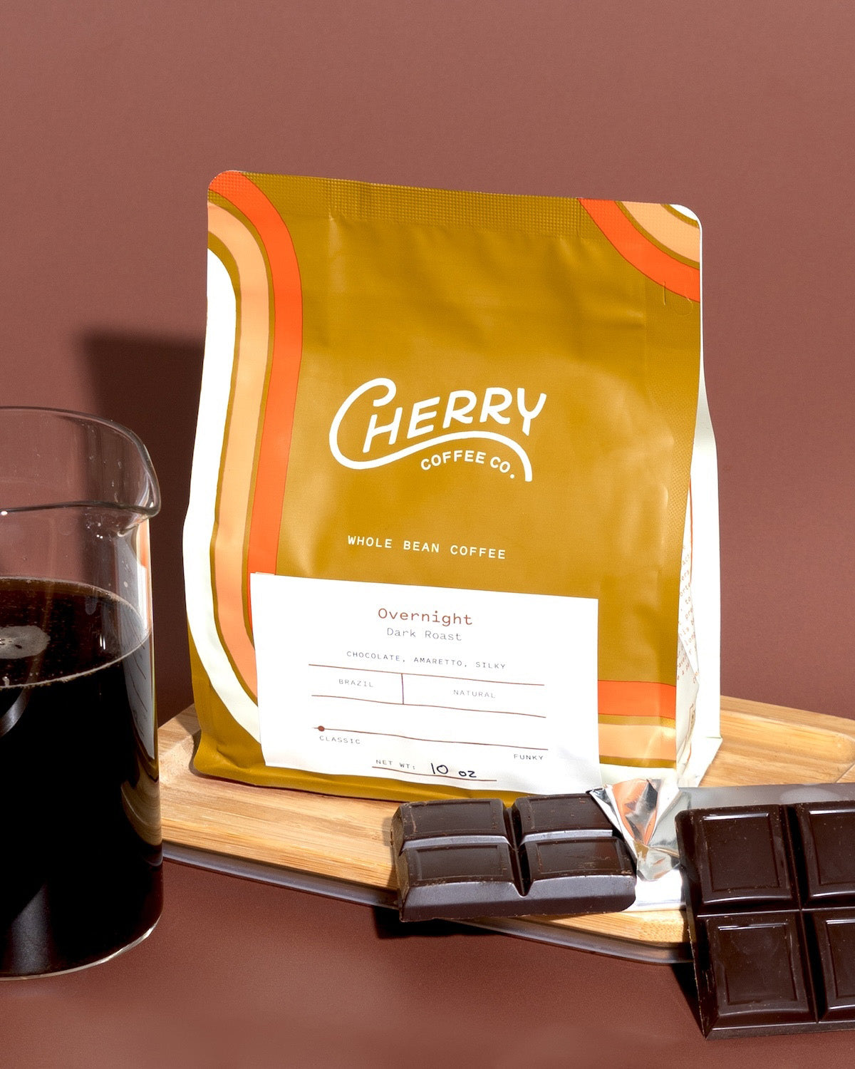 photograph of a 10 oz bag of Overnight dark roast coffee next to a bar of chocolate and carafe of brewed coffee.