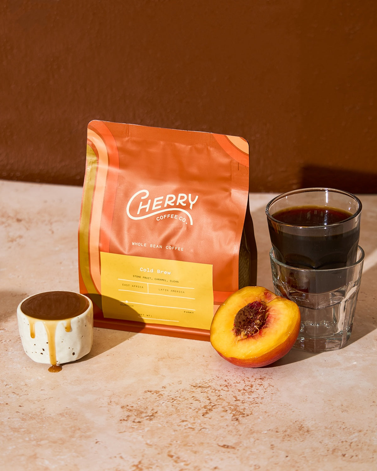 Cherry coffee cold brew retail bag with a small cup of caramel, cold brew, and half of a peach.