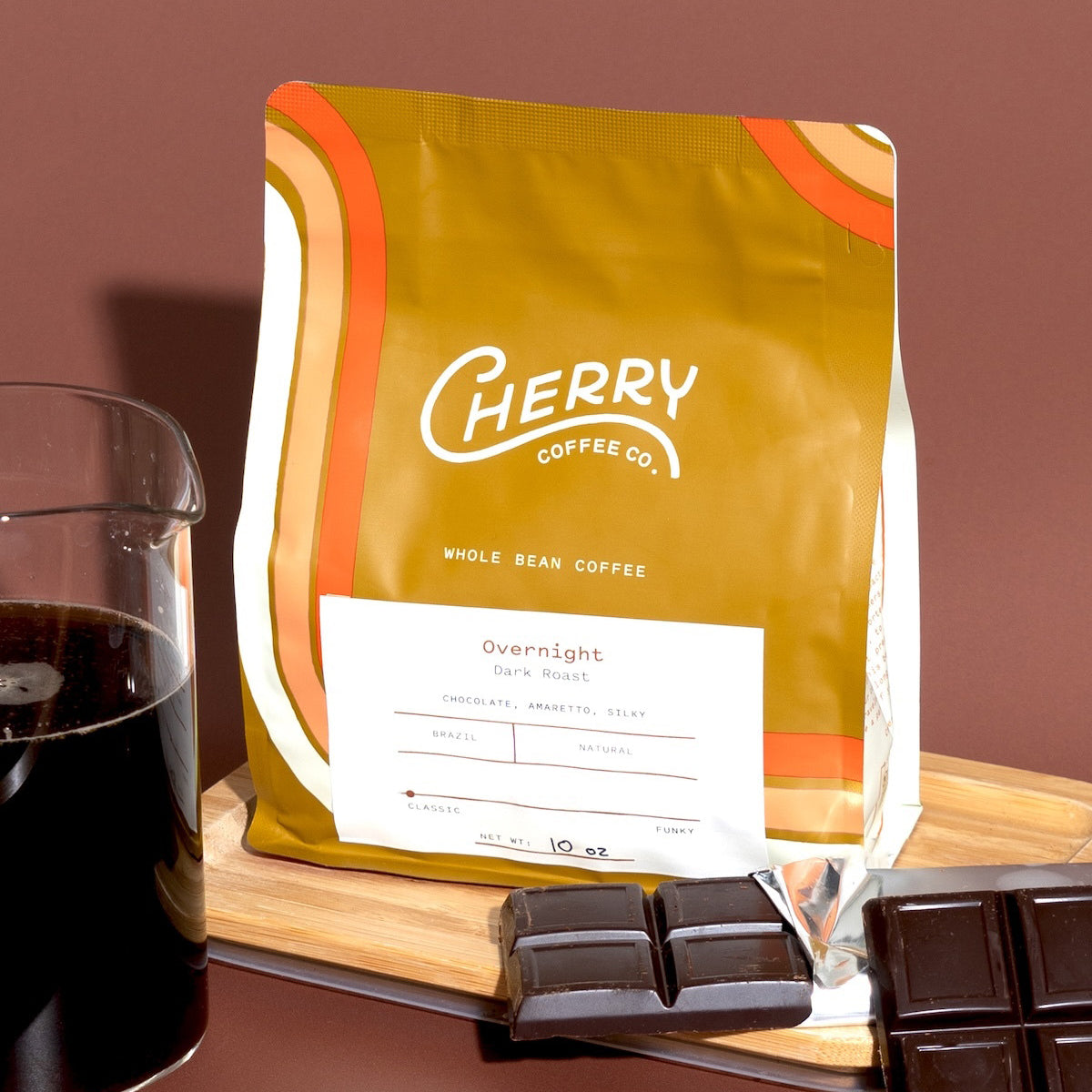 photograph of a 10 oz bag of Overnight dark roast coffee next to a bar of chocolate and carafe of brewed coffee.